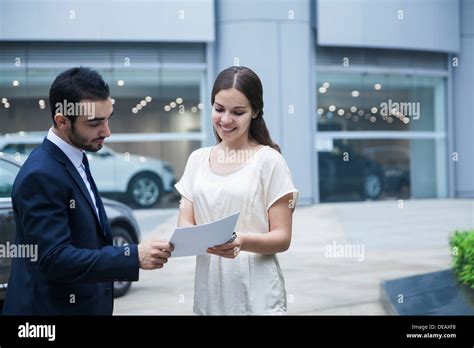 Car Salesman And Young Woman Looking Over The Paperwork At A Car