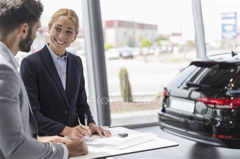 Car Saleswoman And Male Customer Signing Contract Paperwork In Car Dealership Showroom