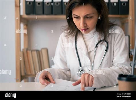 Concentrated Female Doctor Reading Document At Table In Clinic Doctor Cardiologist Doing