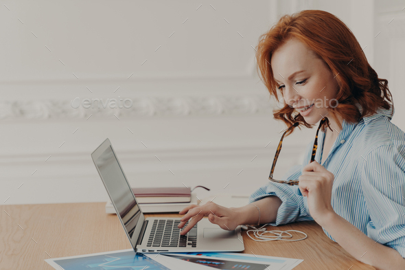 Sideways Shot Of Concentrated Redhead Prosperous Businesswoman Busy With Paperwork Types On